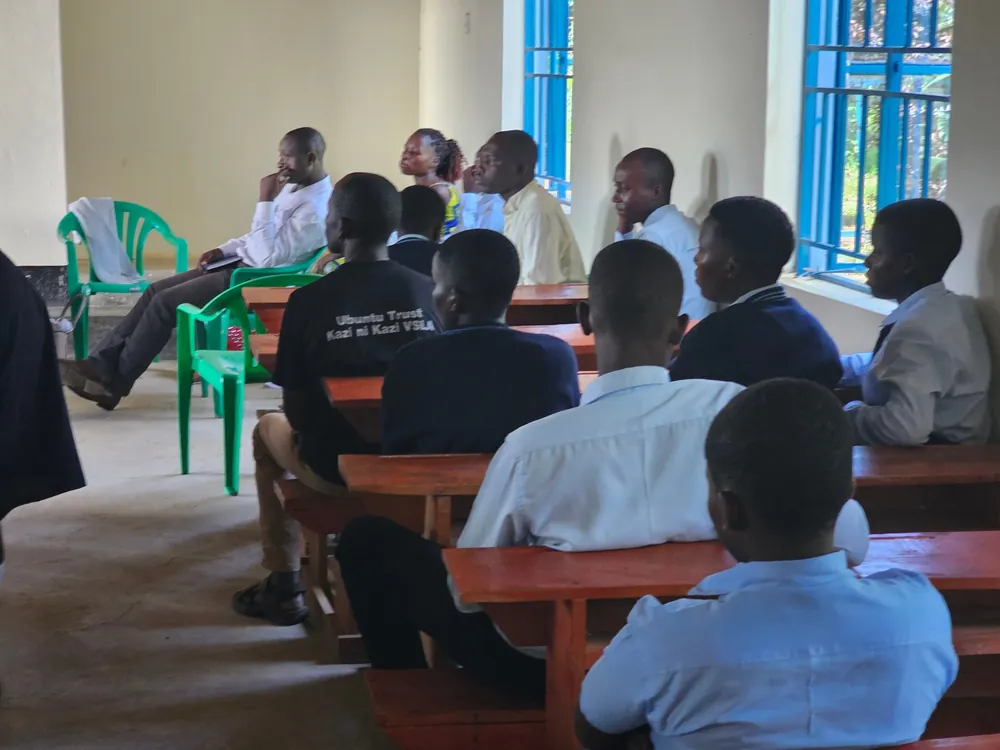 Learners in uniform and adults sit on benches and chairs during a meeting in a classroom.