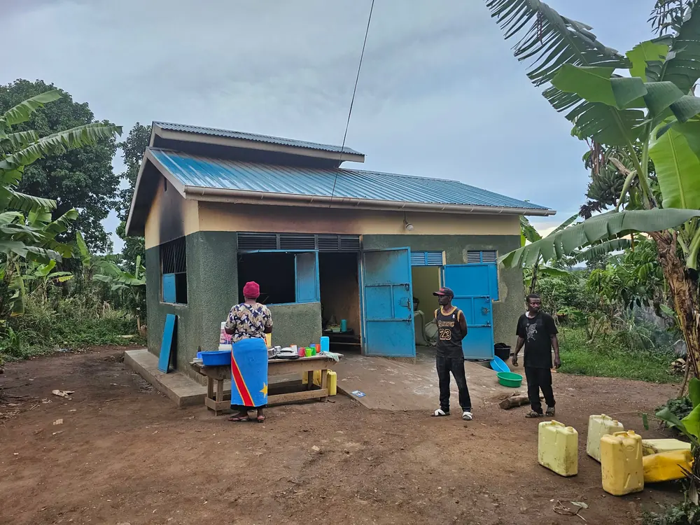 The school kitchen, a small building with a blue roof and blue doors, with a cook preparing food at a table outside and water containers nearby.
