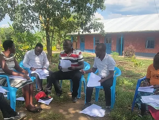 Teachers sit in a circle of plastic chairs under a tree, discussing printed papers outside a school building.