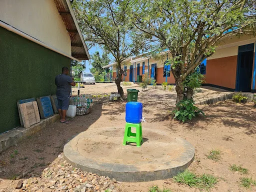 A water container on a green stand at a round concrete water point between school buildings and trees.