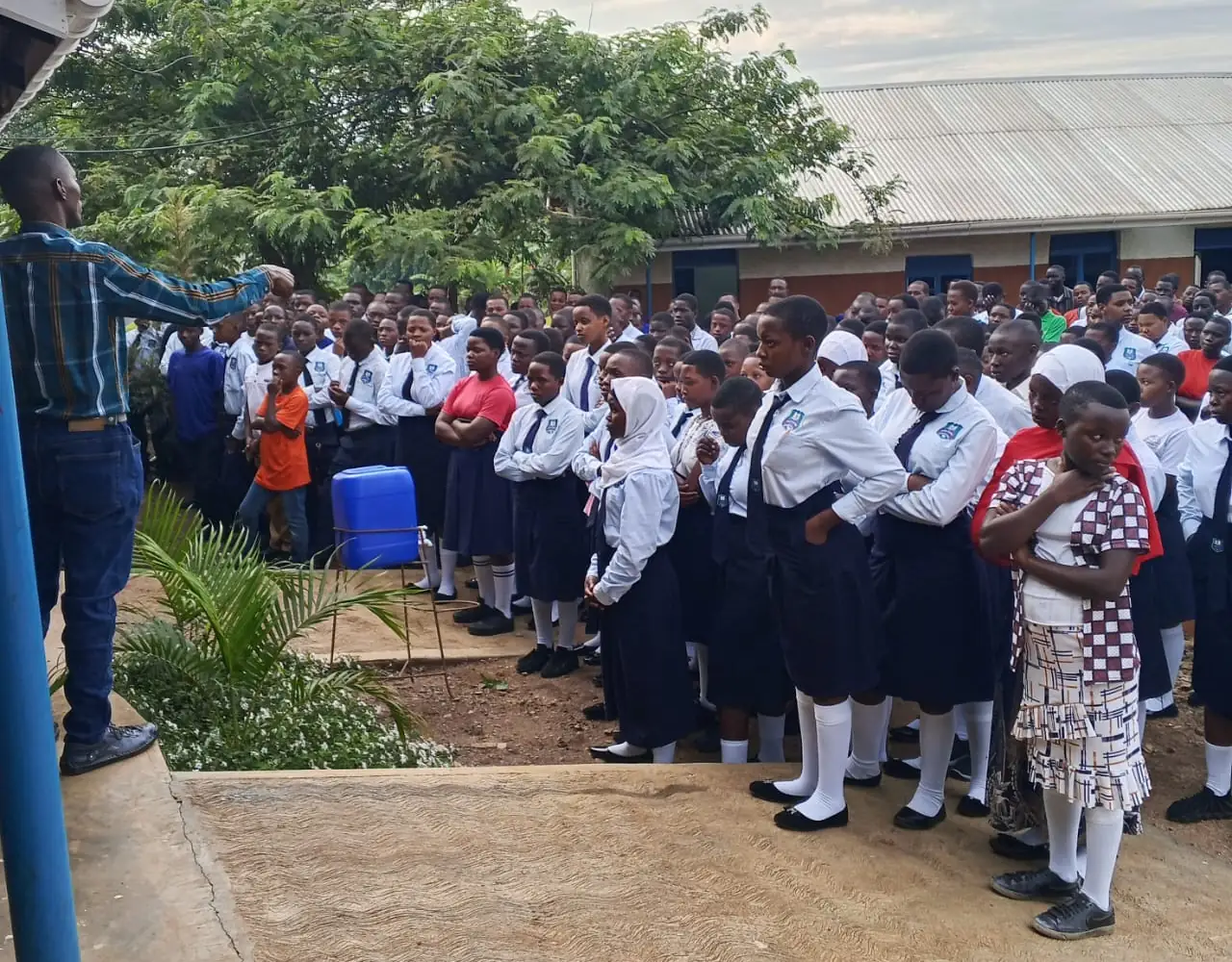 A staff member speaks to a large assembly of learners in school uniform in the school compound.