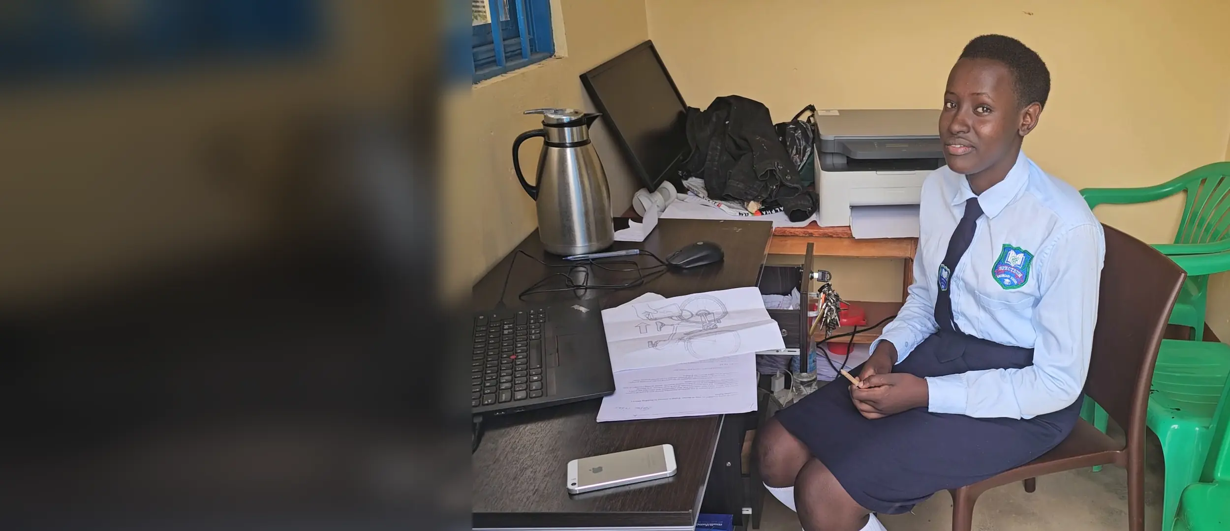 A learner in school uniform sits at a desk beside a laptop and a printed design drawing of a bicycle.