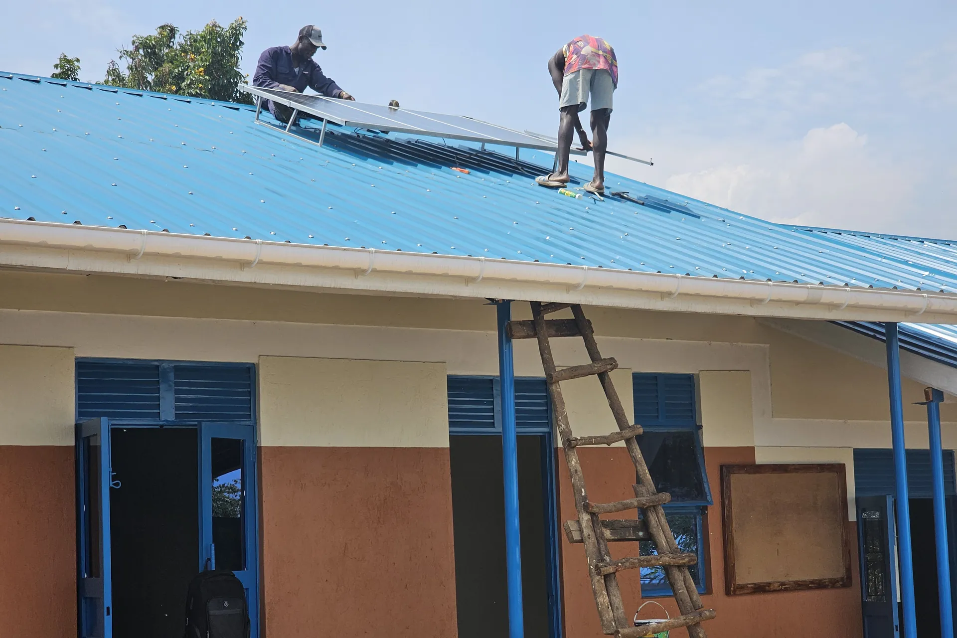Two workers fit solar panels on the blue metal roof of a school building, with a wooden ladder against the wall.